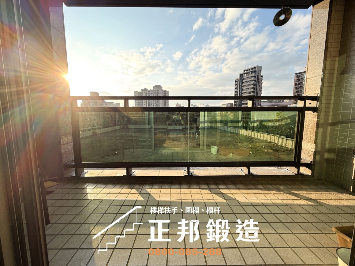 台北松隆路舊大樓陽台欄杆更換後的實景，黑色不鏽鋼烤漆框架與綠半反射強化玻璃沿用原本簡潔分格，遠方可見台北市區建築與天空，陽台地面維持原有磁磚。正邦鍛造以客製化C9F-1015欄杆處理原有金屬鏽蝕問題，並盡量不改變社區原有建築立面風格。
